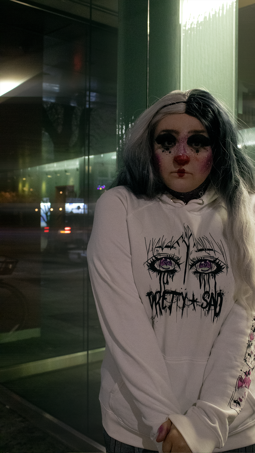 A person in clown makeup wearing a white hoodie with a graphic print of stylized eyes and the text 'Pretty & Sad' on the front.