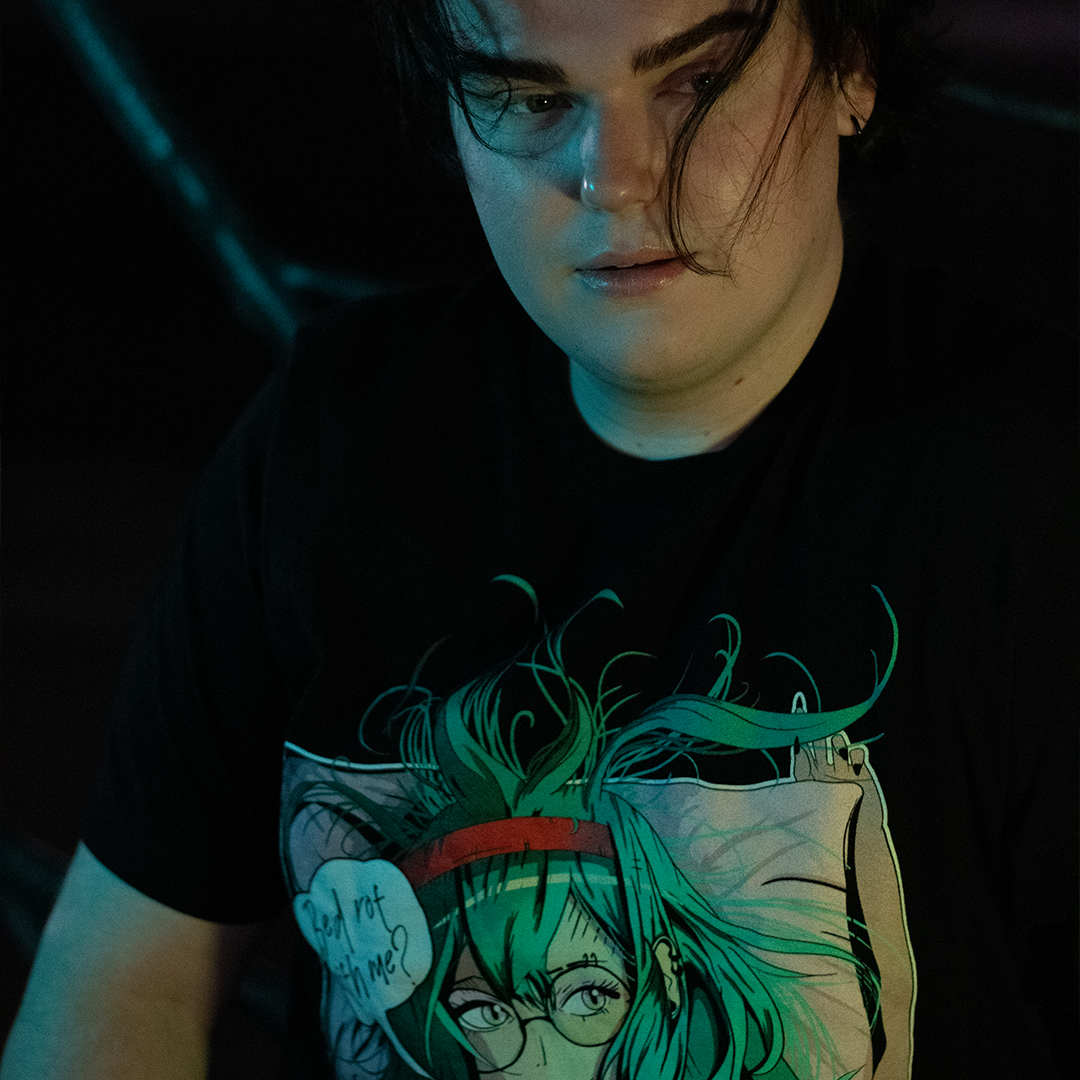 Person in a city wearing black shirt with stylized anime woman with teal hair and bra, speech bubble that says "bed rot with me?"
