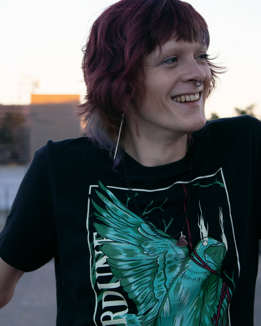 Smiling person wearing black shirt with stylized graphic of a green bird, one wing and neck is wrapped up with a red piece of string. "Therdune" logo written on left side vertically