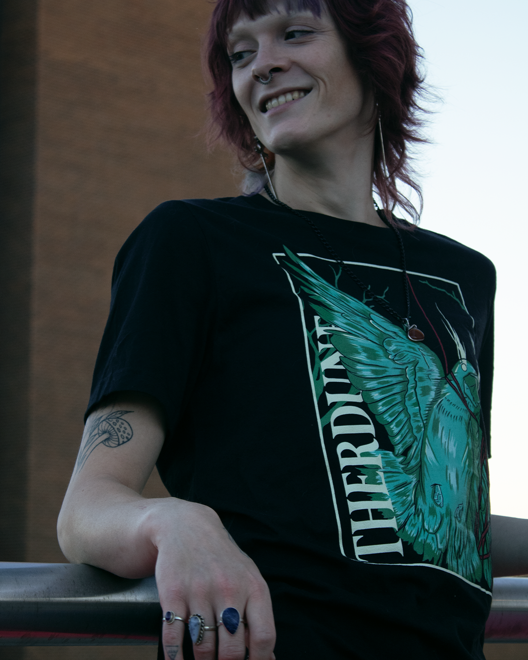 Smiling person wearing a black shirt with stylized graphic of a green bird, one wing and neck is wrapped up with a red piece of string. "Therdune" logo written on left side vertically